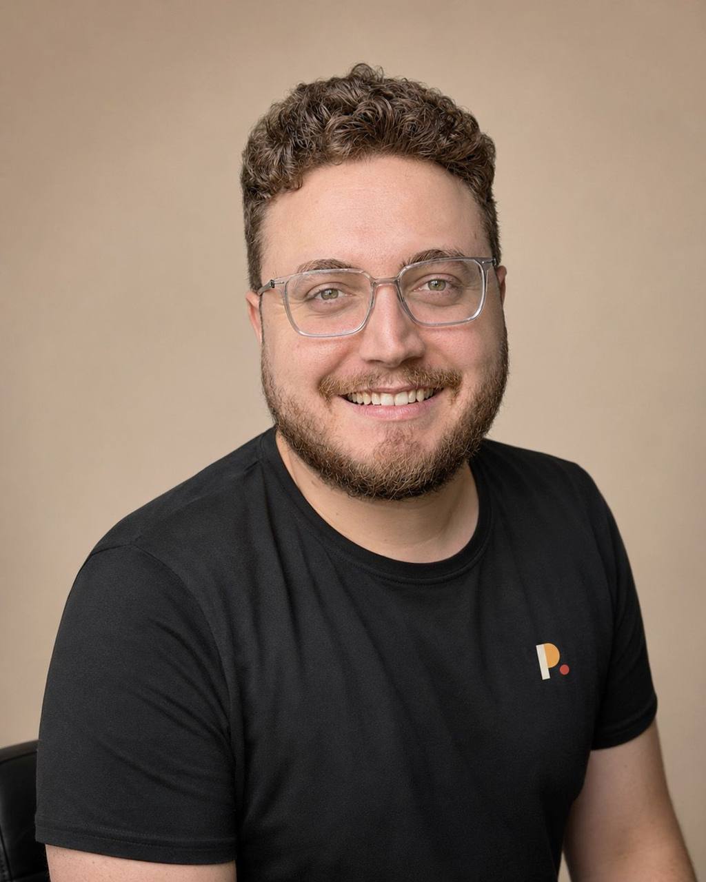 Charles Patoine, souriant avec des lunettes, en t-shirt noir sur fond beige.