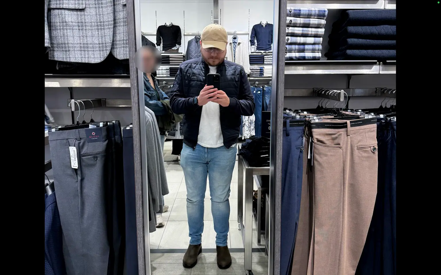 Charles se photographie dans un miroir, en jeans bleus, manteau foncé et casquette beige.