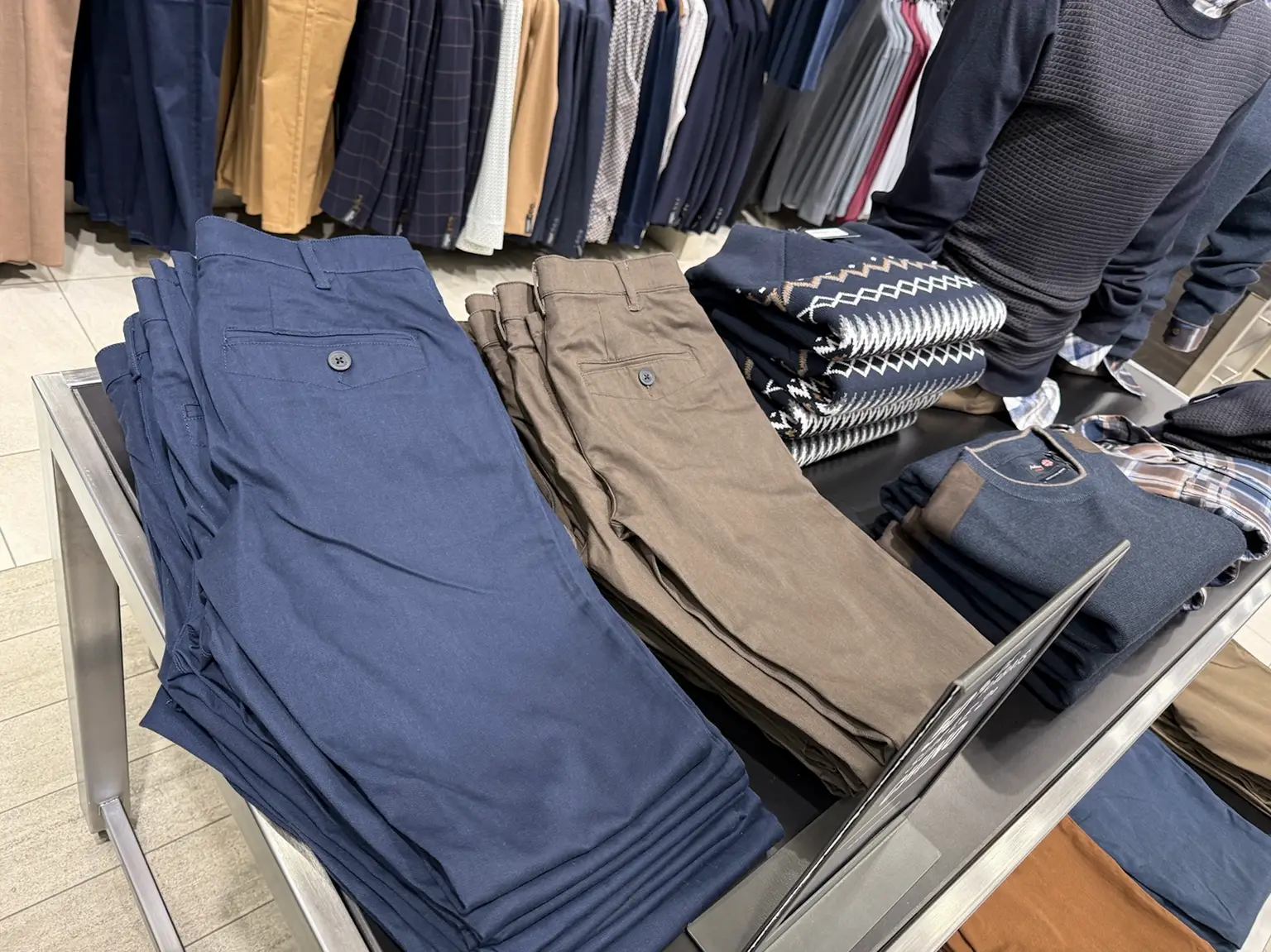 Deux piles de chinos, l’une bleue et l’autre beige taupe, côtoient des chandails sur un présentoir.